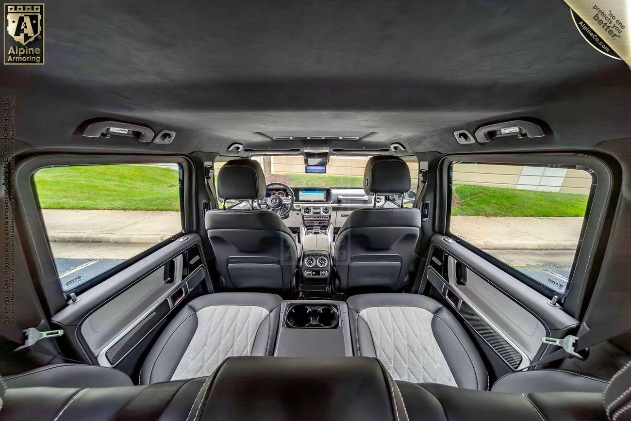 Interior view of a Mercedes-Benz G-Class SUV  showcasing the backseat area with a built-in screen and a rear window covered by a protective panel. The upholstery is dark grey and black.