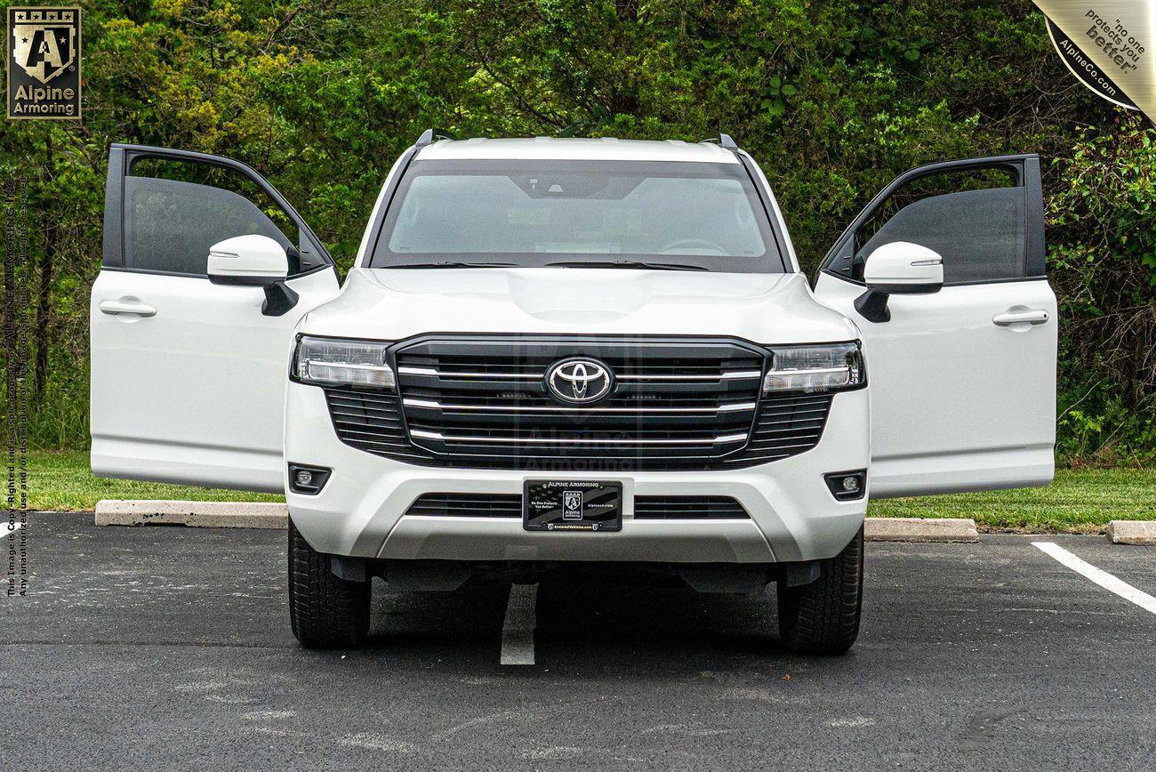 A white armored Toyota LandCruiser 300 SUV with both front doors open, parked outdoors on an asphalt surface near greenery. The vehicle features a prominent front grille and a visible license plate.