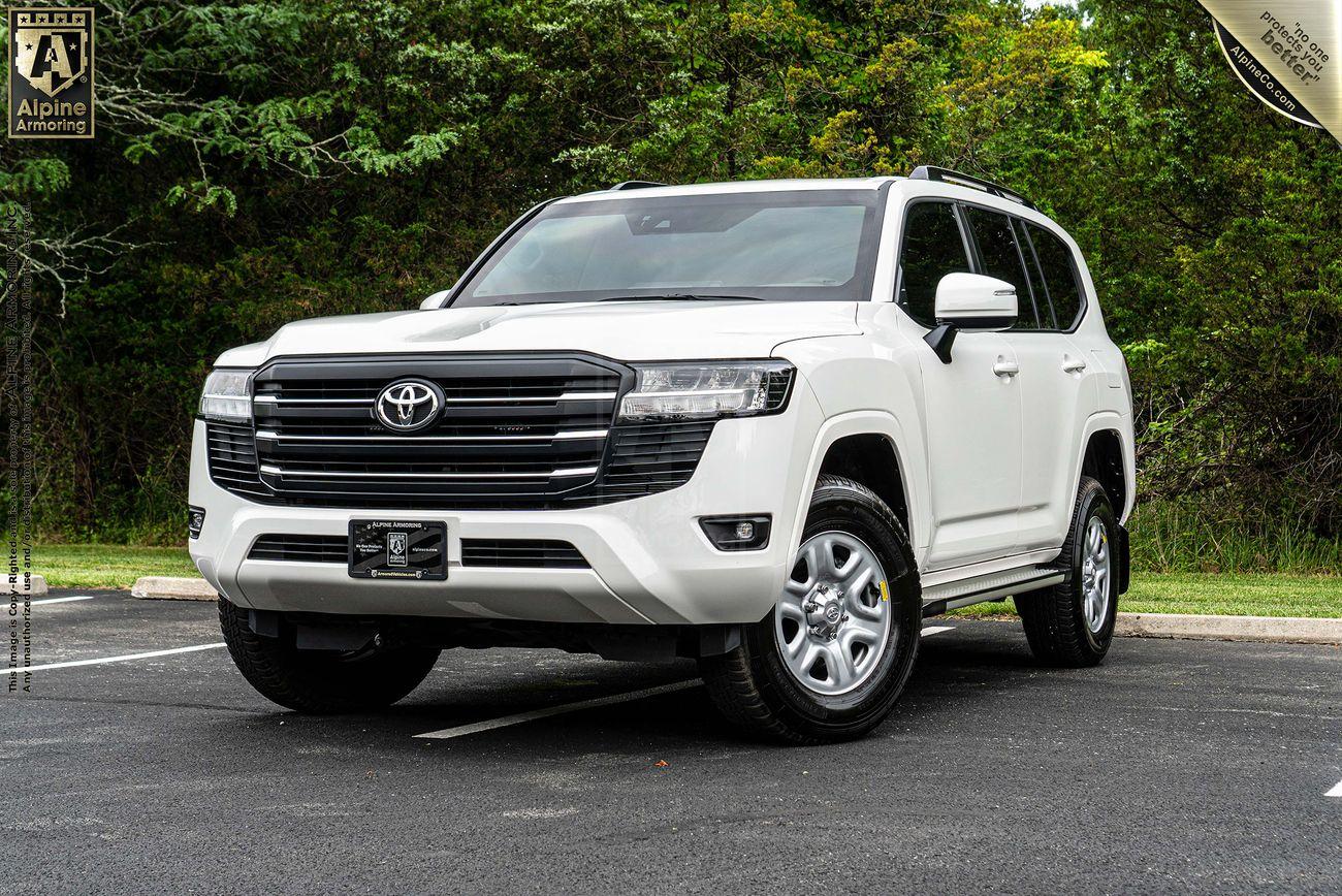 A white armored Toyota LandCruiser 300 SUV is parked in an outdoor setting with trees in the background. The vehicle is positioned slightly angled to the left.