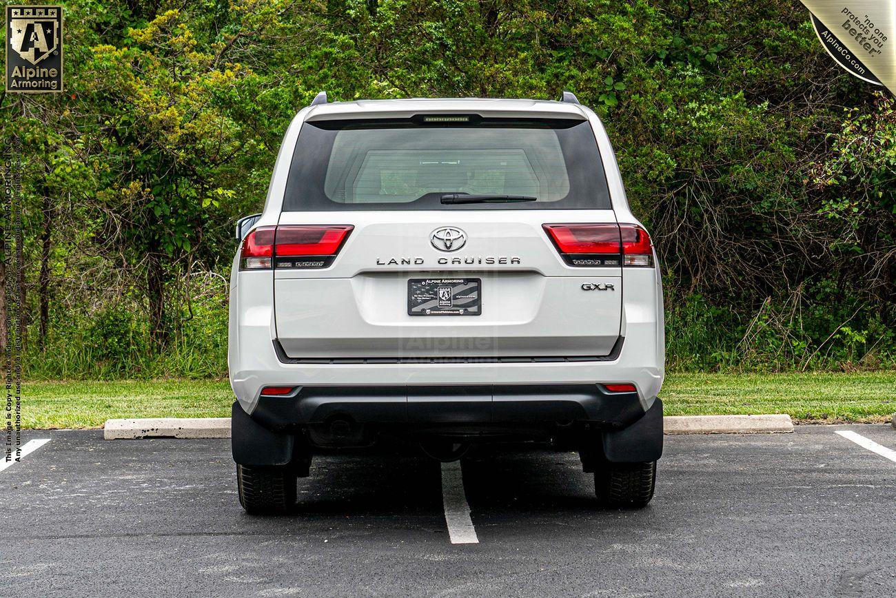 Rear view of a white Toyota Land Cruiser 300 parked in a lot, with surrounding greenery visible in the background. The license plate area has a dealership logo.