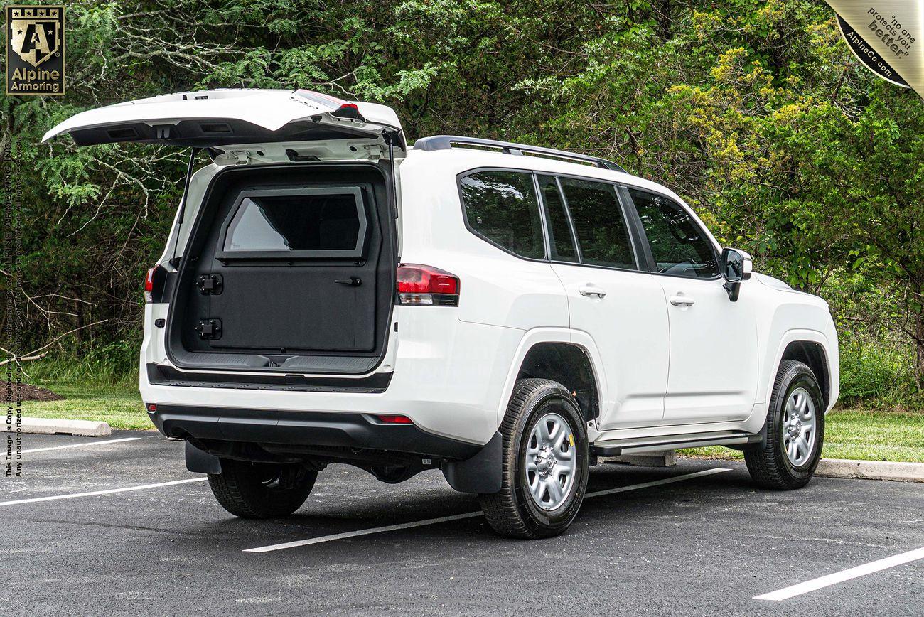 A white armored Toyota LandCruiser 300 SUV is parked in an outdoor lot with its rear hatch open, revealing a secure storage compartment. The vehicle is positioned beside a lush green forest.