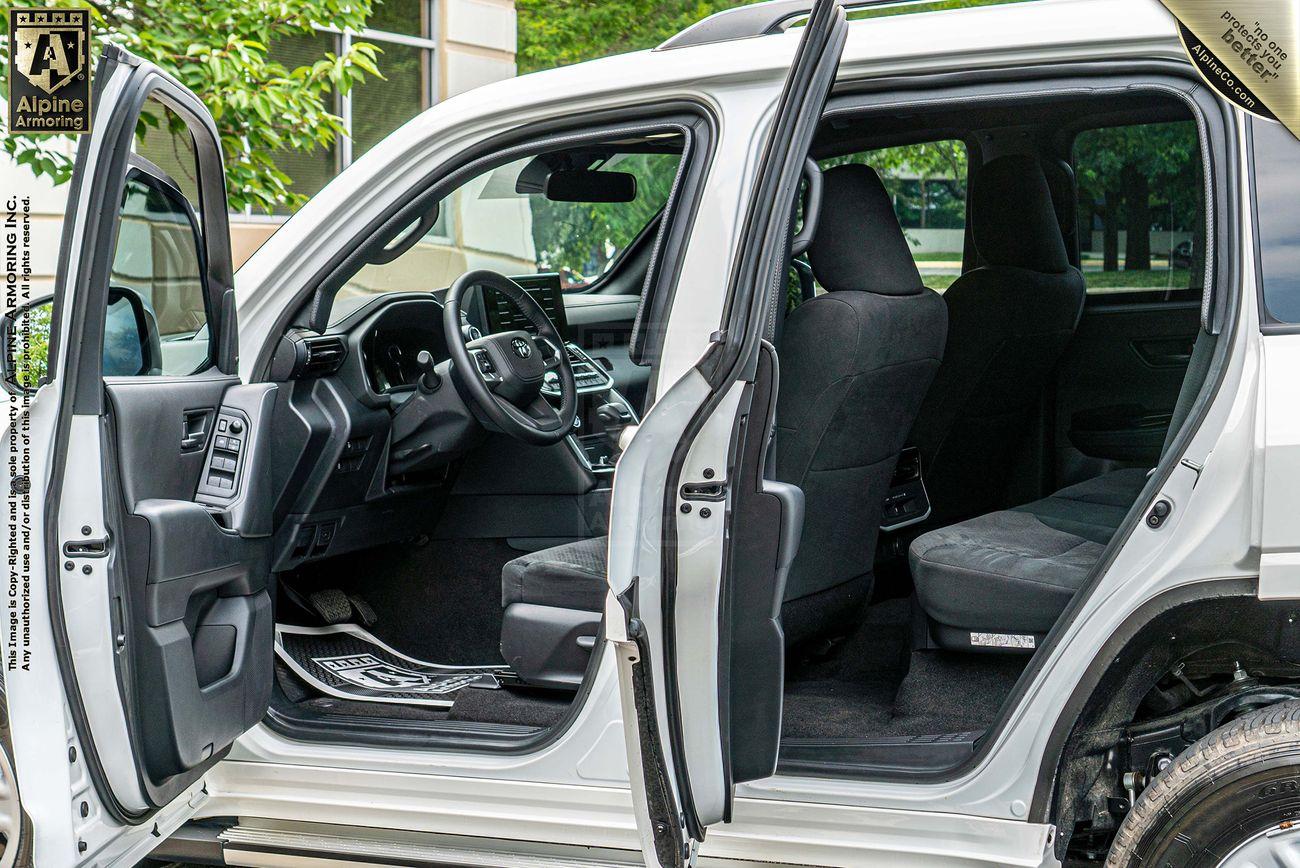 White armored Toyota LandCruiser 300 SUV with front and rear passenger doors open, showing the interior seats, dashboard, and steering wheel. The vehicle is parked next to a building with large windows and greenery outside.