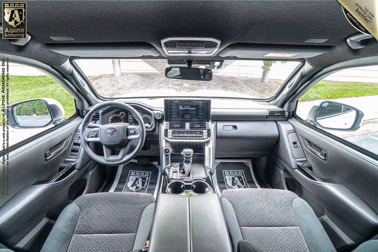 Interior view of an armored Toyota LandCruiser 300 SUV featuring a touchscreen dashboard display, steering wheel with controls, and various amenities. The seats are upholstered in gray fabric. The logo "Alpine Armoring" is visible.