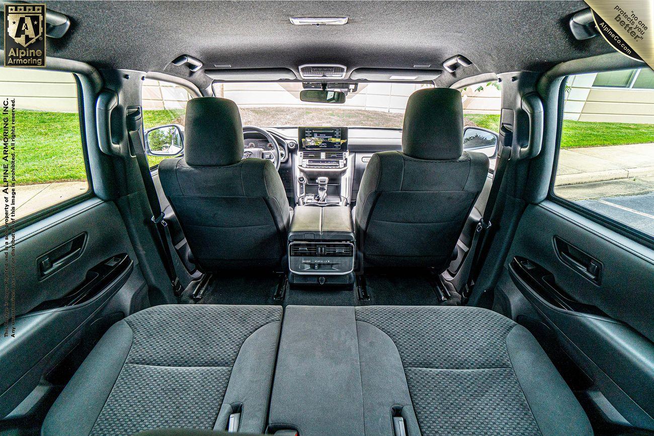 Interior of an armored Toyota LandCruiser 300 viewed from the back seat, showing the front seats, center console, and dashboard with an Alpine Armoring logo in the top left corner.