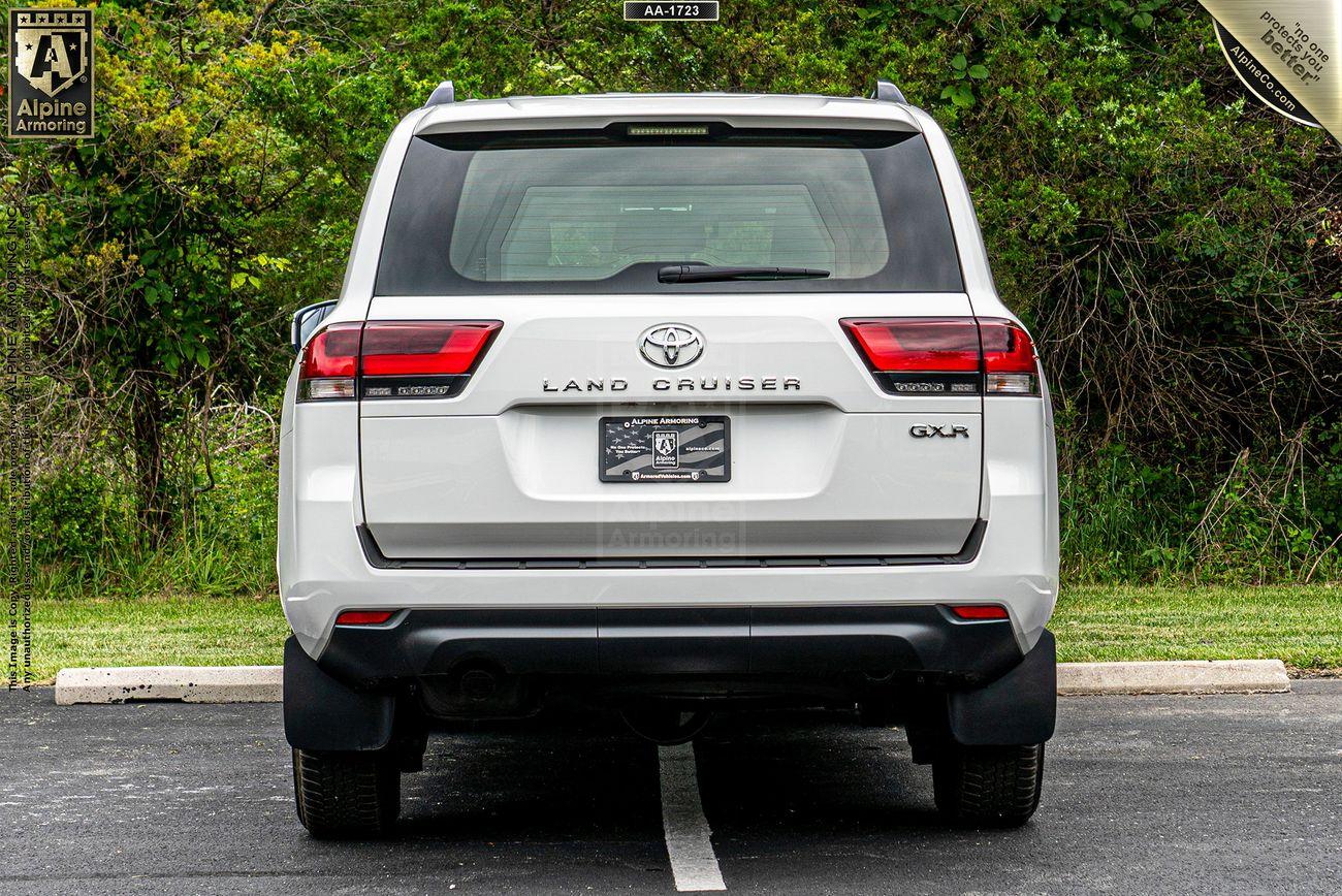 Rear view of a white armored Toyota Land Cruiser 300 SUV parked in a lot with trees and foliage in the background. License plate area blurred.