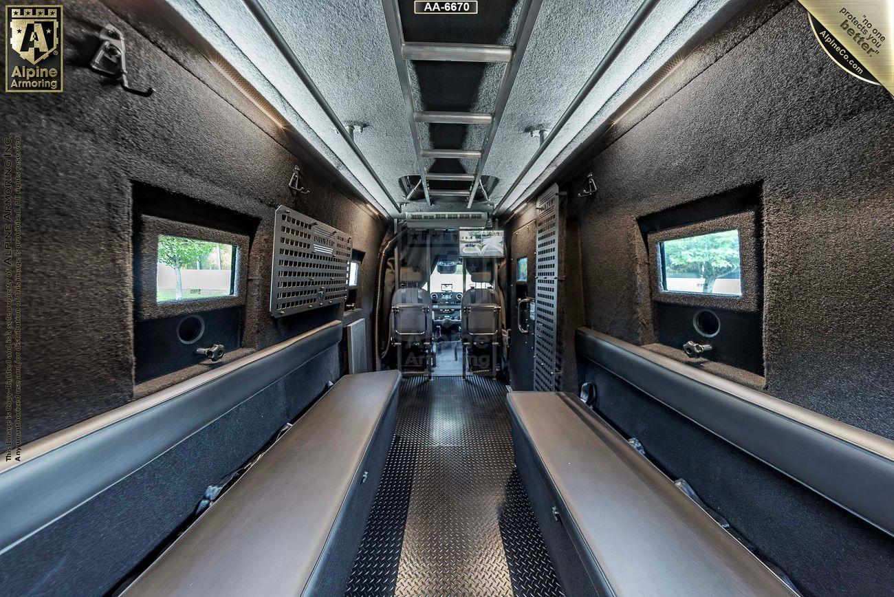 Interior of an armored SWAT Pointer Van from Alpine Armoring, featuring two bench seats along the walls, small windows, and metal flooring, with equipment storage areas on one side.