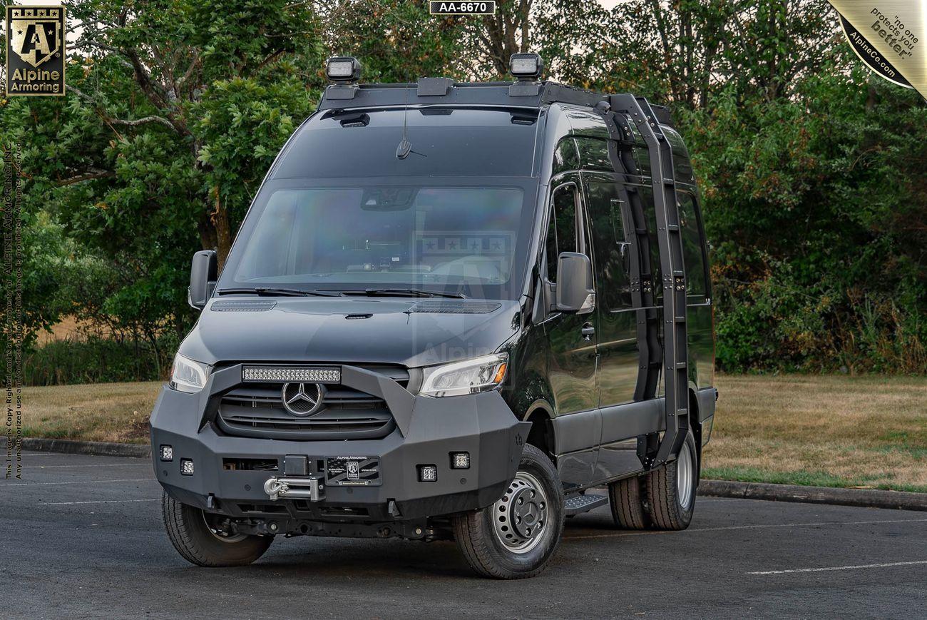 A black armored SWAT Pointer Van from Alpine Armoring is parked in an outdoor area with trees in the background. The vehicle displays the Alpine Armoring logo and has various equipment mounted on the roof.