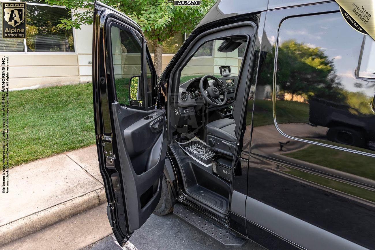 The driver's side door of a black  SWAT Pointer Van from Alpine Armoring is open, revealing a modern interior with steering wheel and controls visible. The car is parked on a street, with a grassy area and building in the background.