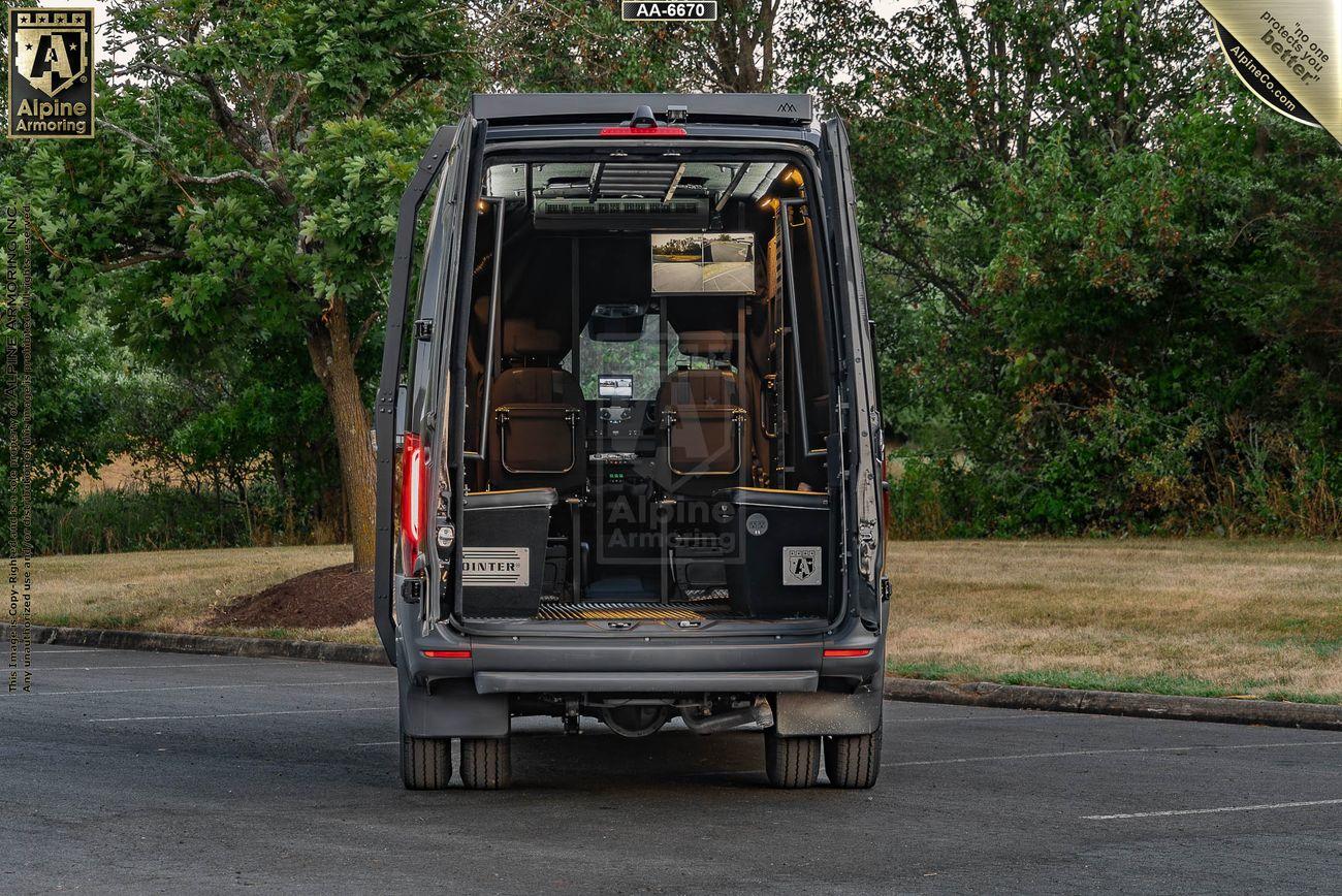 A SWAT Pointer Van from Alpine Armoring with its rear doors open, revealing an empty interior and a single seat front-facing inside. The van is parked in an outdoor area with trees and grass in the background.