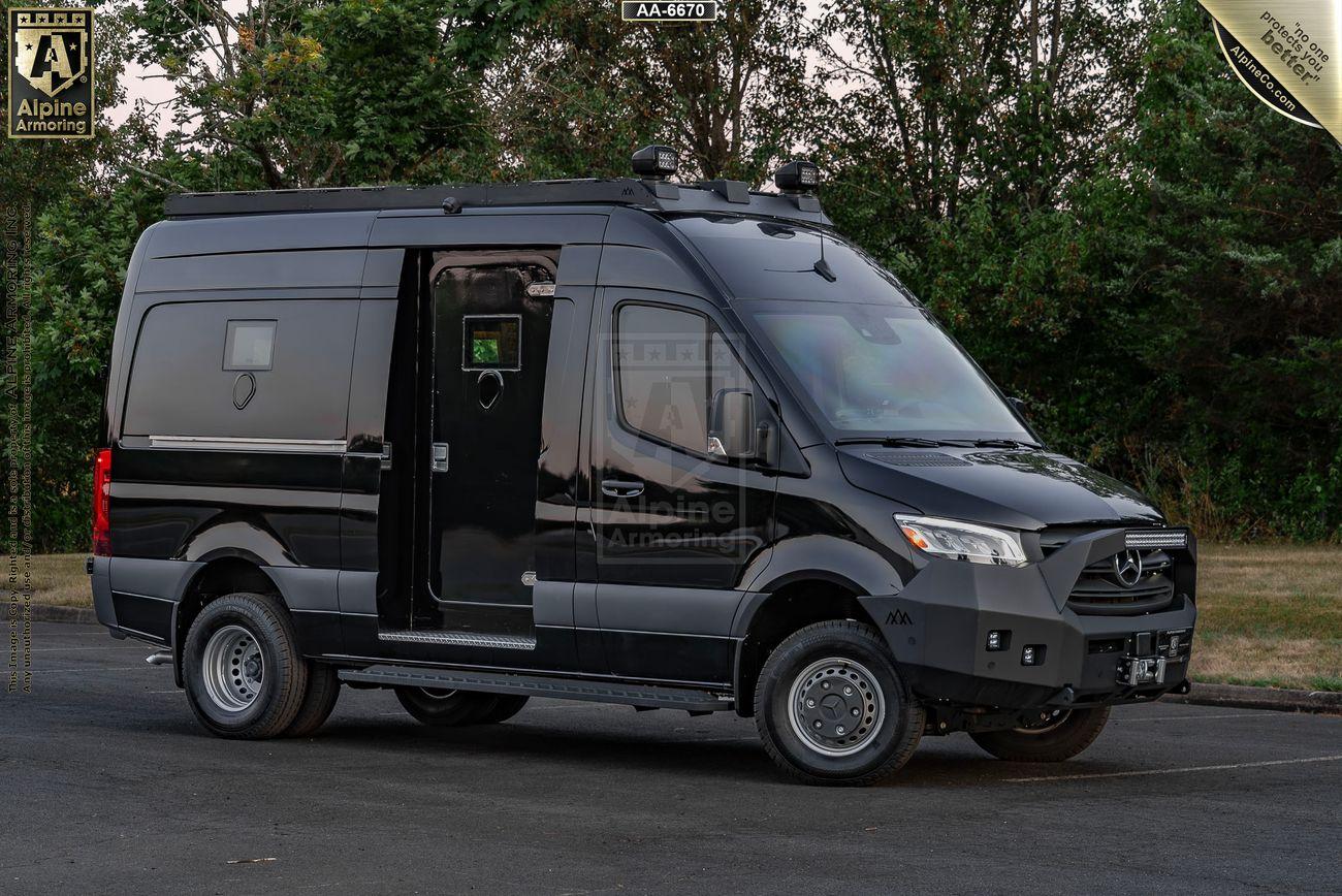 A black, SWAT Pointer Van from Alpine Armoring parked on a paved surface with trees in the background. The van has additional features and equipment visible on its exterior.