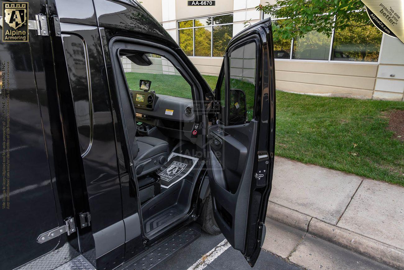 A black SWAT Pointer Van from Alpine Armoring is parked on the street with its driver's side door open, revealing the interior. The van is positioned next to a grass patch and a building.