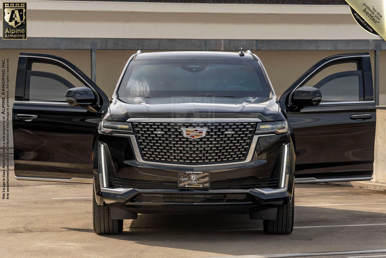 A black bulletproof Cadillac Escalade SUV with both front doors open is parked in front of a building. The vehicle has a distinctive front grille and is positioned centrally in the image.