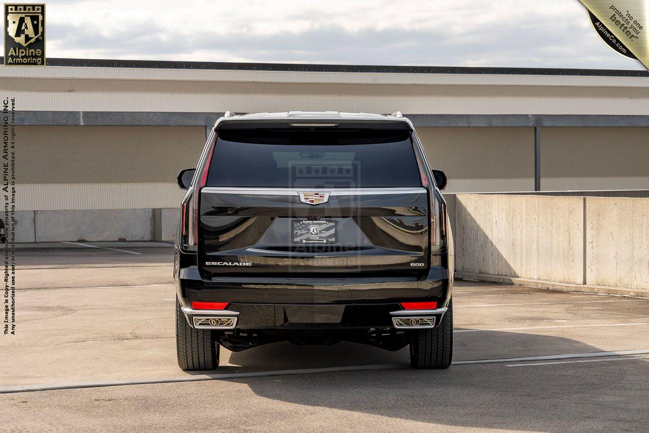 A rear view of a black armored Cadillac Escalade SUV parked in an outdoor lot, featuring a license plate holder with a logo and a concrete wall in the background.