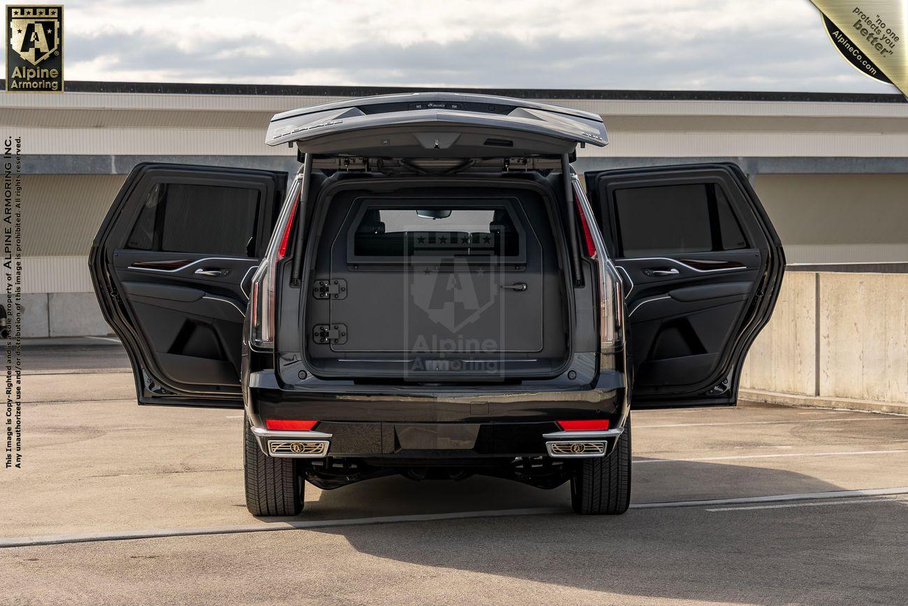 A black bulletproof Cadillac Escalade SUV with its rear doors fully open, showing an empty rear cargo area. The vehicle is parked on a rooftop parking area with a concrete barrier and a cloudy sky in the background.