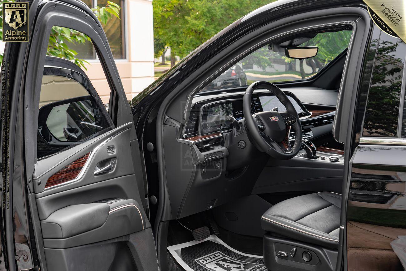 Interior of a bulletproof Cadillac Escalade SUV, showing the driver's seat, steering wheel, and dashboard. The driver's door is open, revealing a sleek design with leather and wood accents.