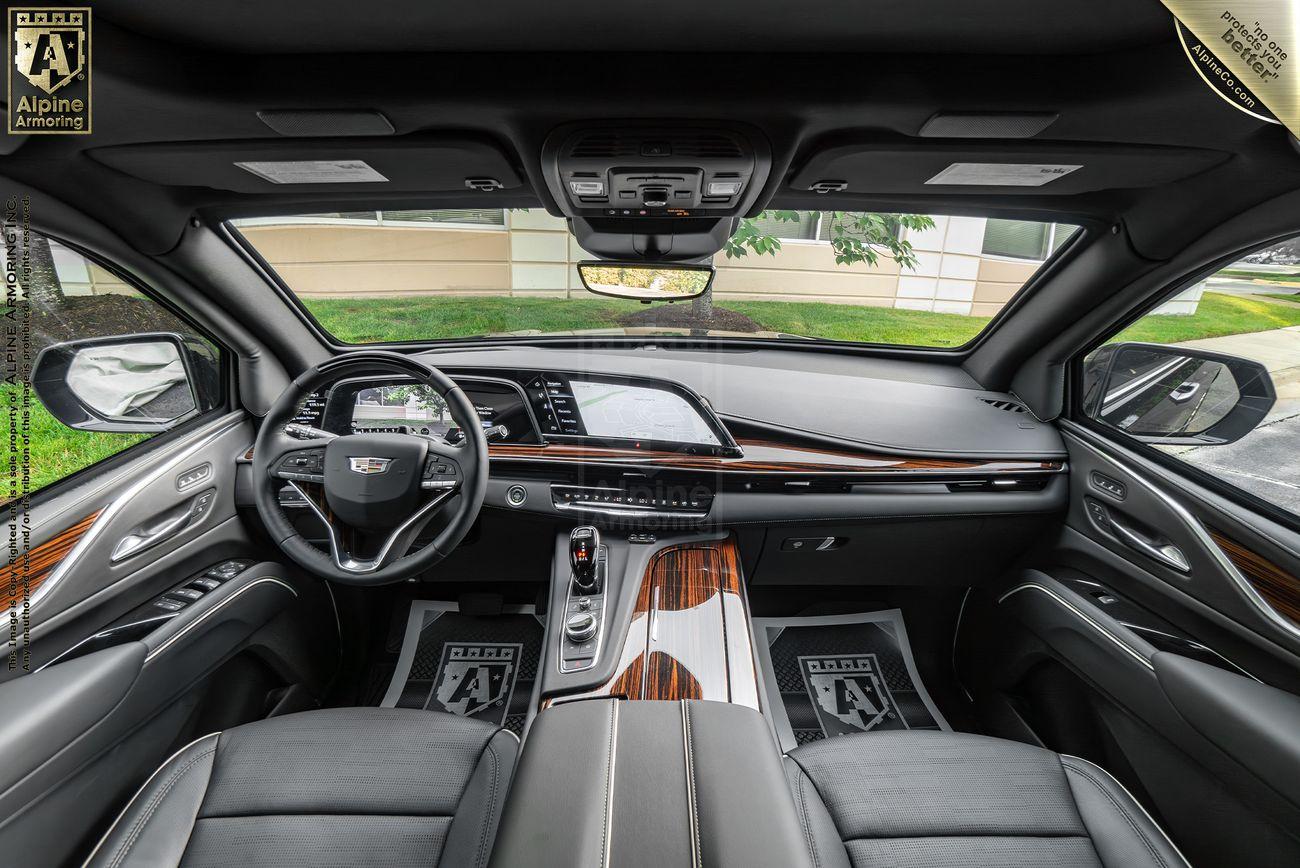 An armored Cadillac Escalade SUV's interior featuring a high-tech dashboard with a large touchscreen, leather seats, and wood trim. The steering wheel has multiple controls and the windshield shows a view outside.