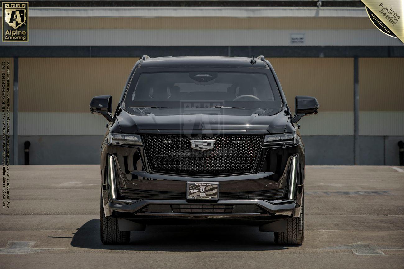 Front view of a black armored Cadillac Escalade ESV SUV parked on a concrete surface in front of a building. The vehicle features distinctive vertical headlights and a prominent grille with the Cadillac logo.