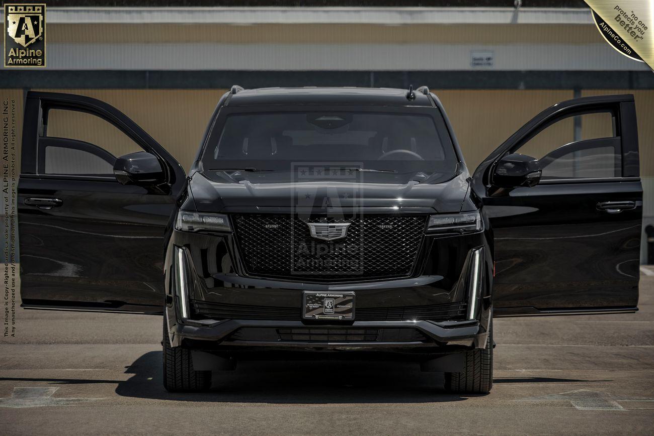 Front view of a black armored Cadillac Cadillac Escalade ESV SUV with both front doors open, parked in a garage with a "Alpine Armoring" sign above.