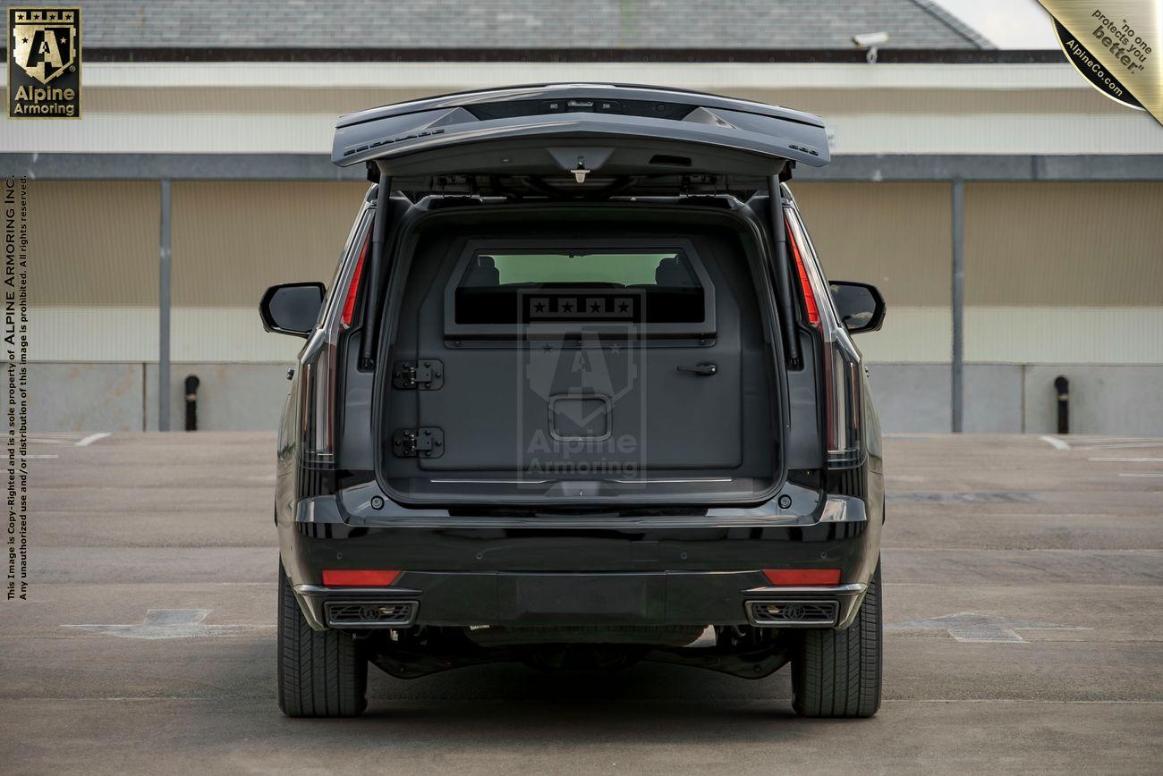 A black Cadillac Escalade ESV SUV with its rear hatch open, parked in an empty lot. The logo "Alpine Armoring" is visible on the inside of the vehicle.