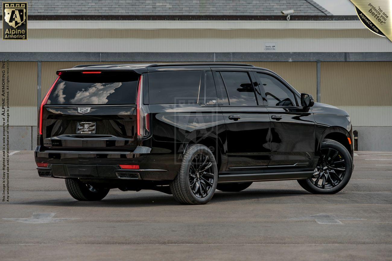 A black bulletproof Cadillac Escalade ESV SUV is parked on a paved surface near a building. The vehicle has tinted windows and black rims.