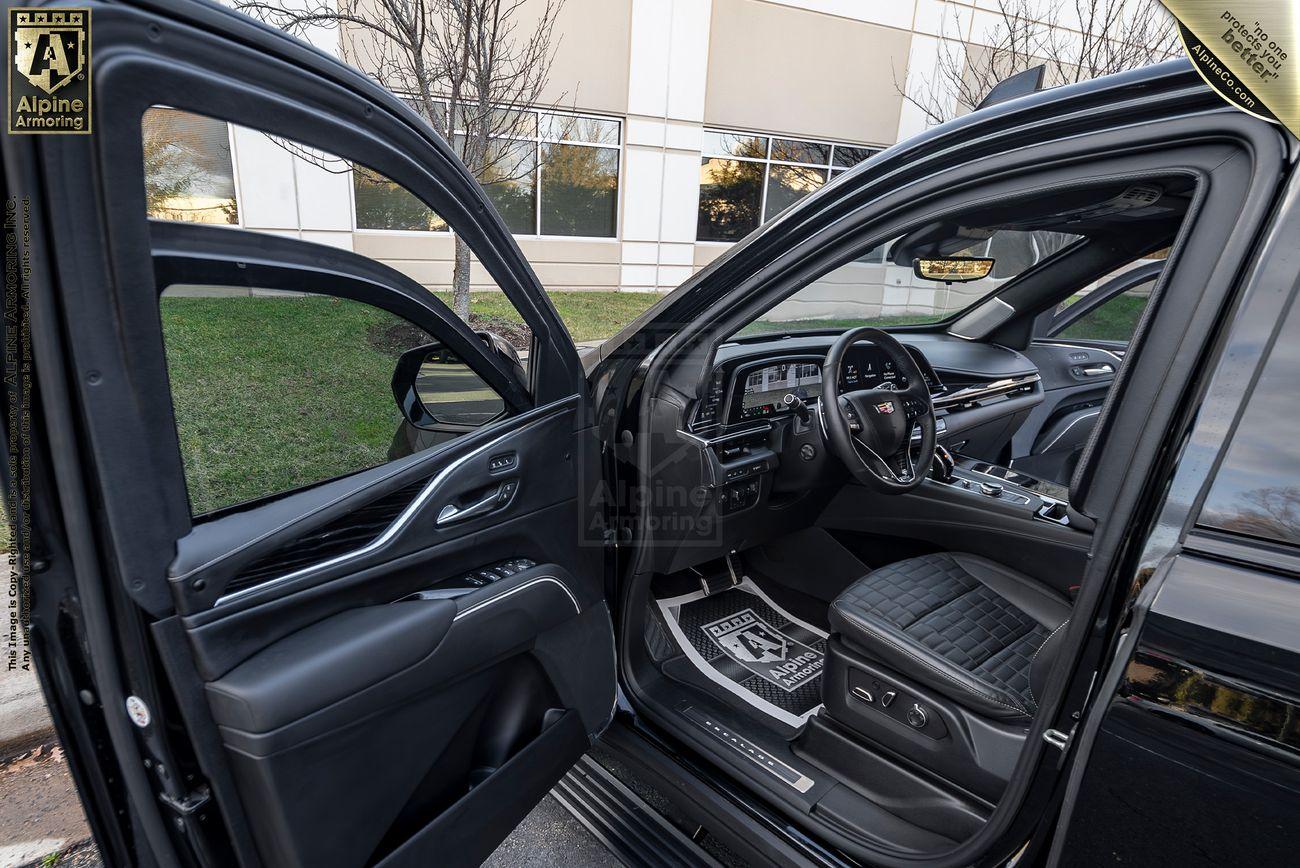 Interior view of an armored black luxury Cadillac Escalade ESV SUVwith both the front and rear driver-side doors open, showcasing the driver's seat, dashboard, and detailed upholstery. Trees and a building are in the background.