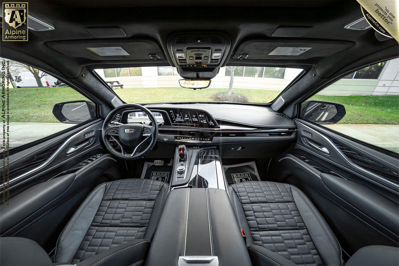 Interior of a Cadillac Escalade ESV SUV cabin with black leather seats, a modern dashboard, and a central console. The image is taken from the back seat, showcasing the steering wheel and infotainment screens.