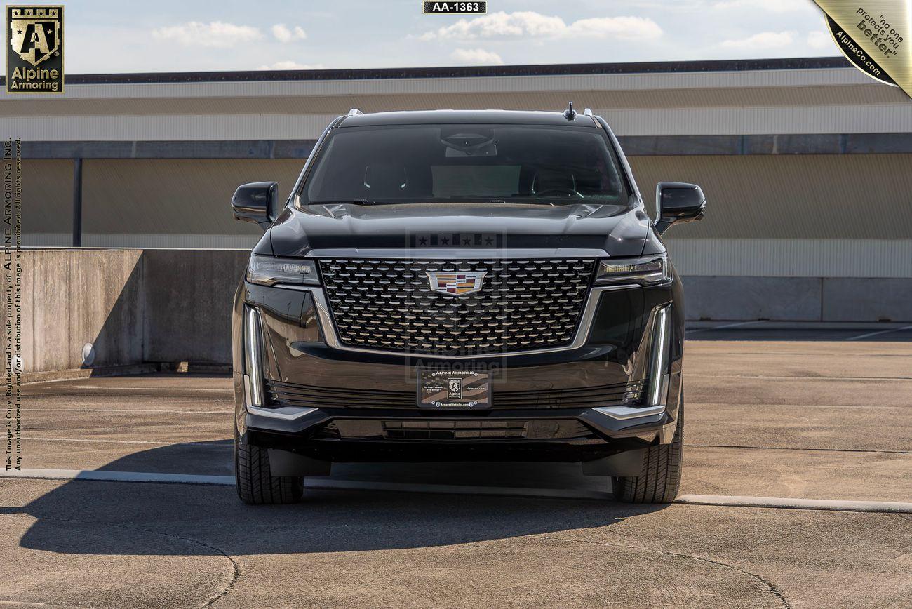 Front view of an armored black Cadillac Escalade ESV Luxury SUV in an outdoor setting, parked on a concrete surface with a building and blue sky in the background.