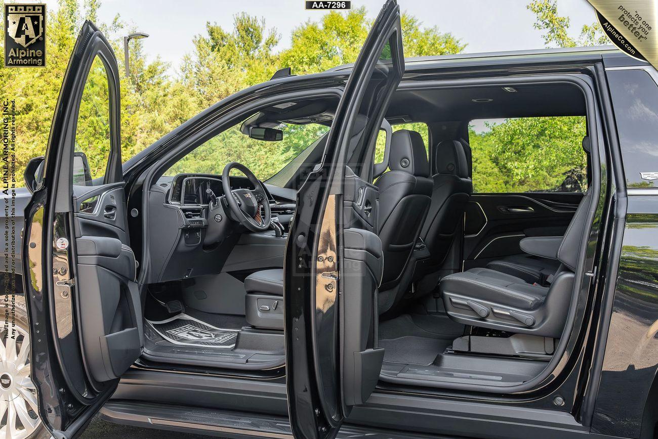 An armored Cadillac Escalade ESV Luxury SUV with front and rear passenger doors open, showcasing a spacious interior with black leather seats and modern dashboard. The vehicle is parked outdoors with green foliage in the background.