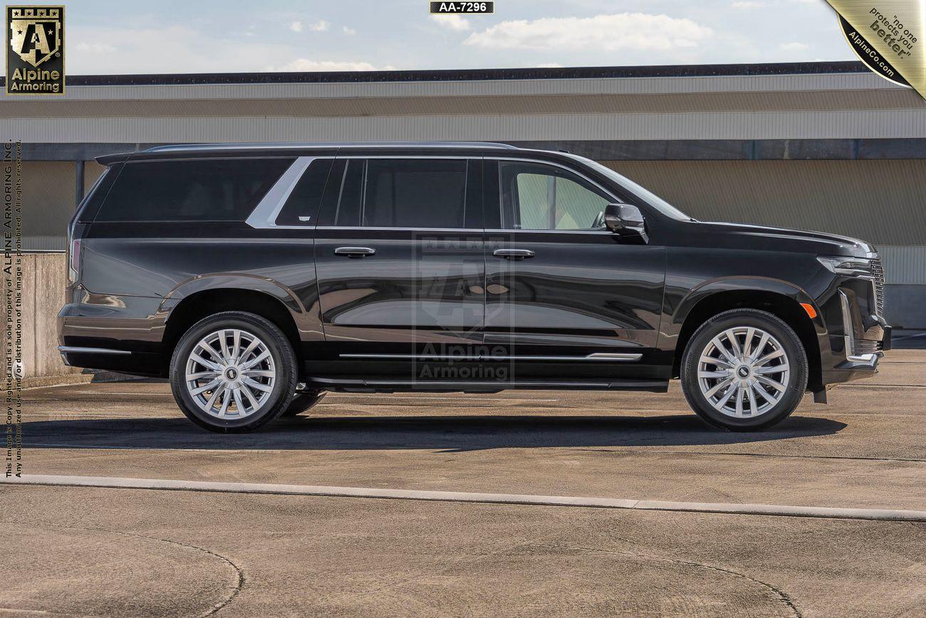 A black armored Cadillac Escalade ESV Luxury SUV with white wheels, parked on a paved surface in front of a building. The vehicle features Alpine Armoring branding.