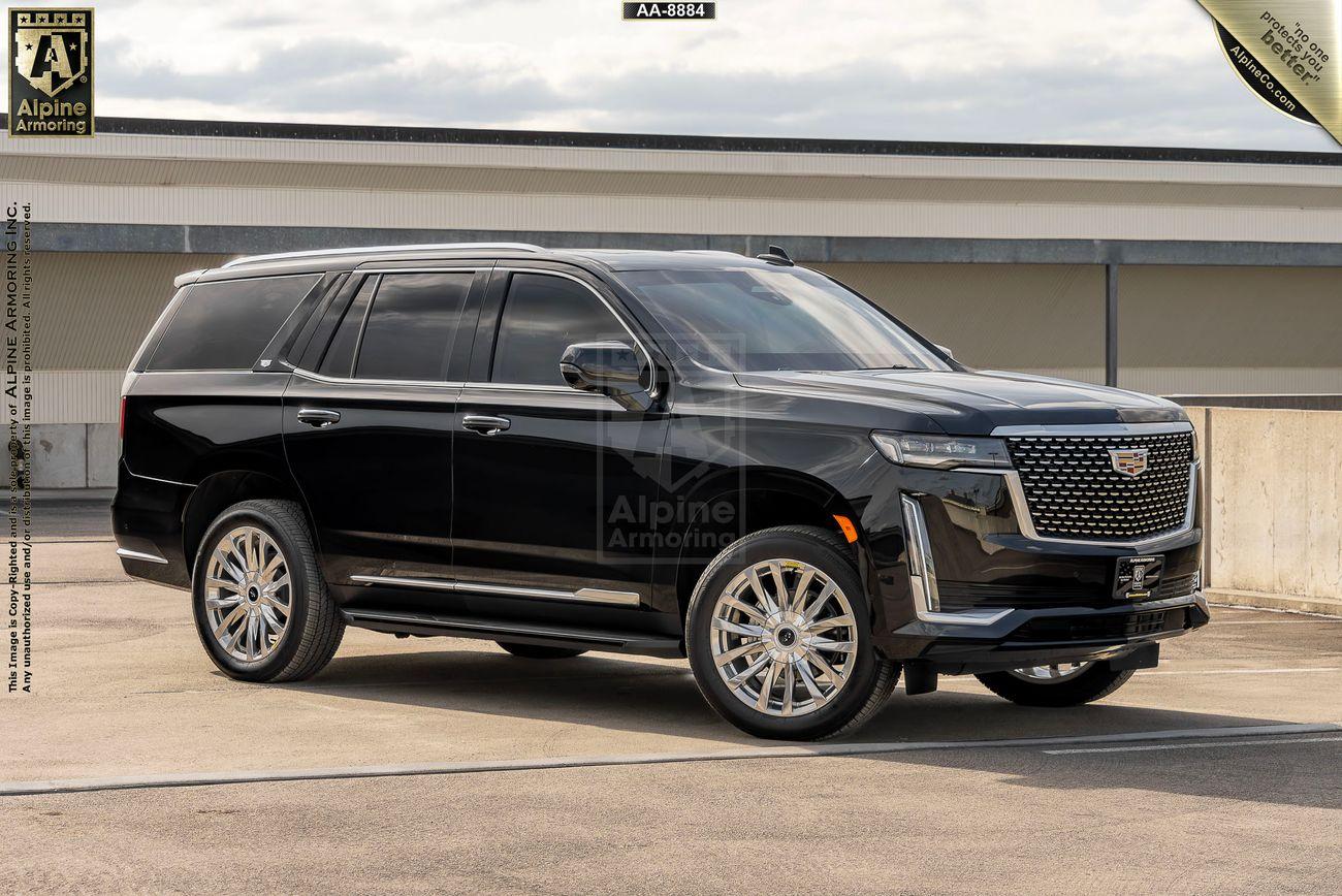 A black, armored Cadillac Escalade Premium Luxury SUV with a polished exterior is parked in an outdoor area. The vehicle features a prominent grille and large wheels. Logos for Alpine Armoring are visible on the sides and front.