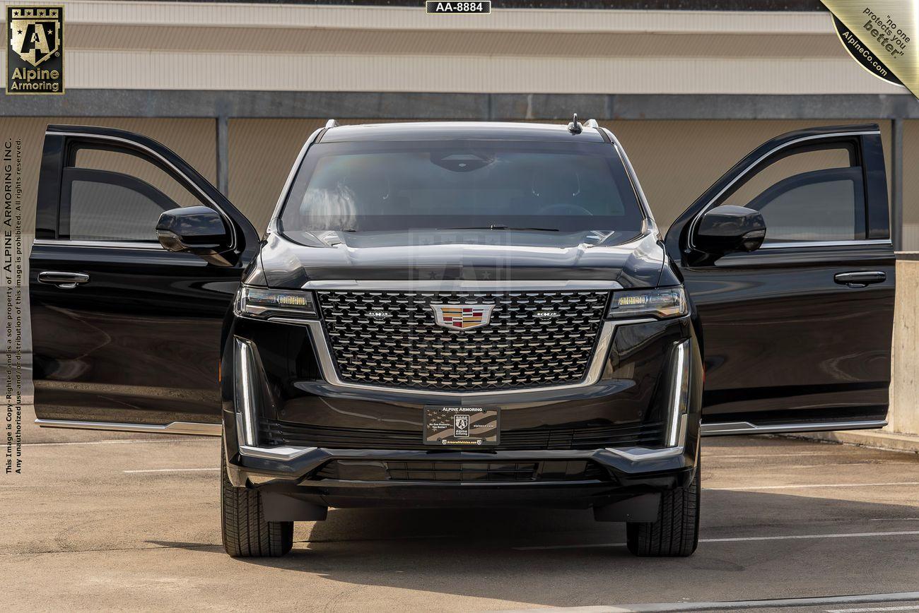 A black armored Cadillac Escalade Premium Luxury SUV with its front doors open, parked on a concrete surface in front of a building. A logo and text are visible in the top left corner.