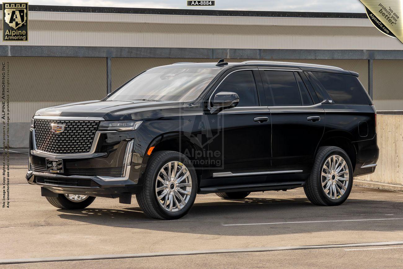 A black Cadillac Escalade Premium Luxury SUV is parked on a rooftop. It has a modern design with large alloy wheels and tinted windows. A banner in the background displays a logo and text.