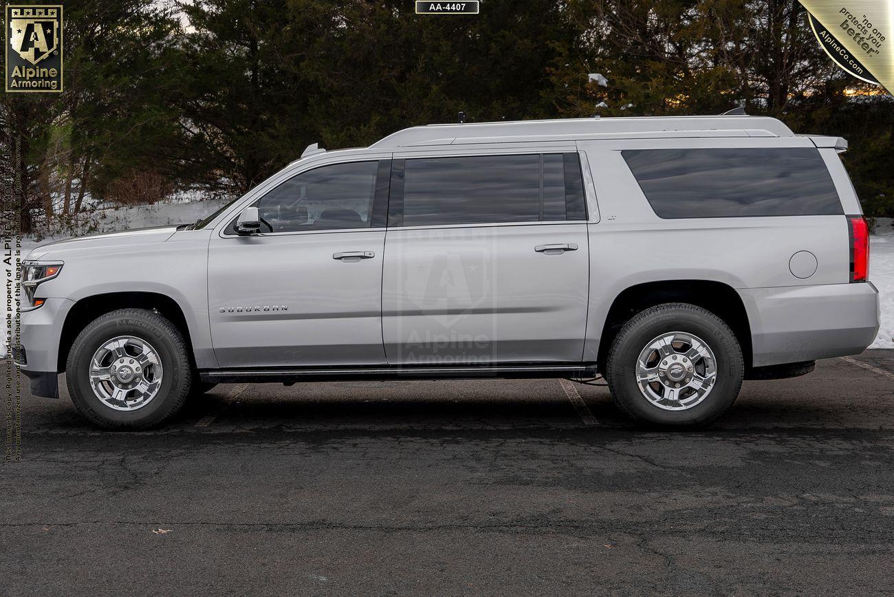 Side view of a white, full-sized armored Chevrolet Suburban 3500HD LT SUV parked on a paved surface with trees in the background.