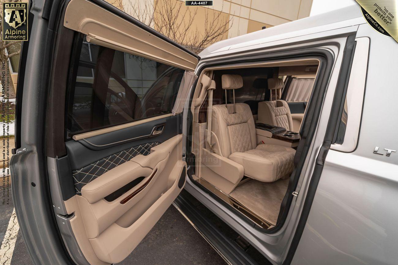 An open door of a Chevrolet Suburban 3500HD LT VIP Limo revealing a spacious, beige leather interior with cushioned seats and an armrest compartment. The exterior of the building is partially visible in the background.