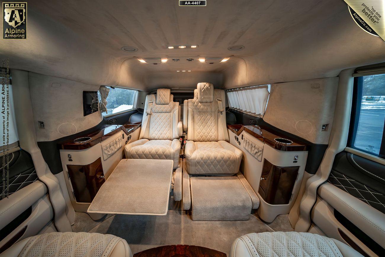 The interior of a luxurious bulletproof Chevrolet Suburban 3500HD LT VIP Limo with cream-colored leather seats, wood accents, and a central table. The vehicle features ample legroom, ambient lighting, and high-end finishes.