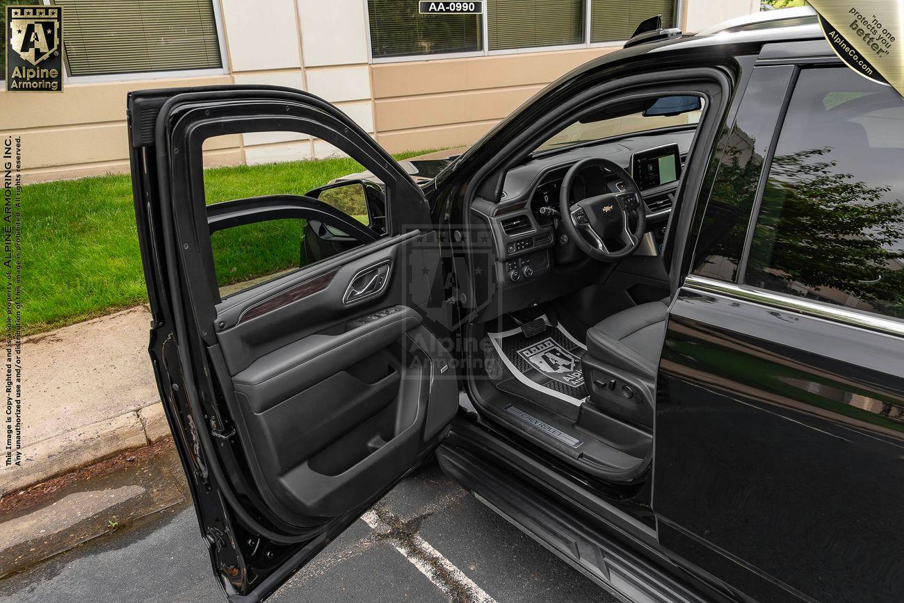 Open driver-side door of a black armored Chevrolet Suburban LT SUV, revealing a black interior with accessible driver's seat, steering wheel, and dashboard. The vehicle is parked with a building and grass visible in the background.