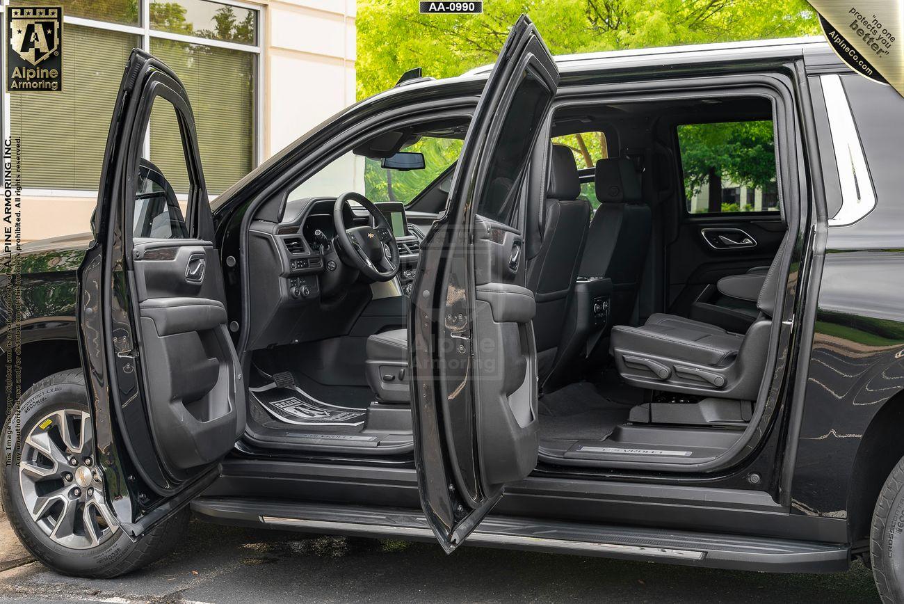 A black armored Chevrolet Suburban LT SUV with all four doors open, showcasing the interior with black leather seats and a spacious cabin. The exterior setting includes trees and part of a building.