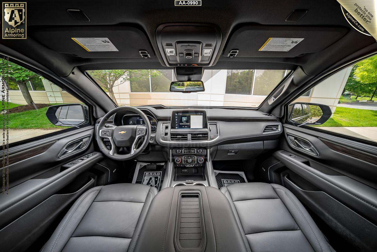 Interior view of an armored Chevrolet Suburban LT SUV cabin featuring a Chevrolet logo on the steering wheel, a central touchscreen, and black leather seats. Panoramic windows show an outdoor scene with trees and buildings.