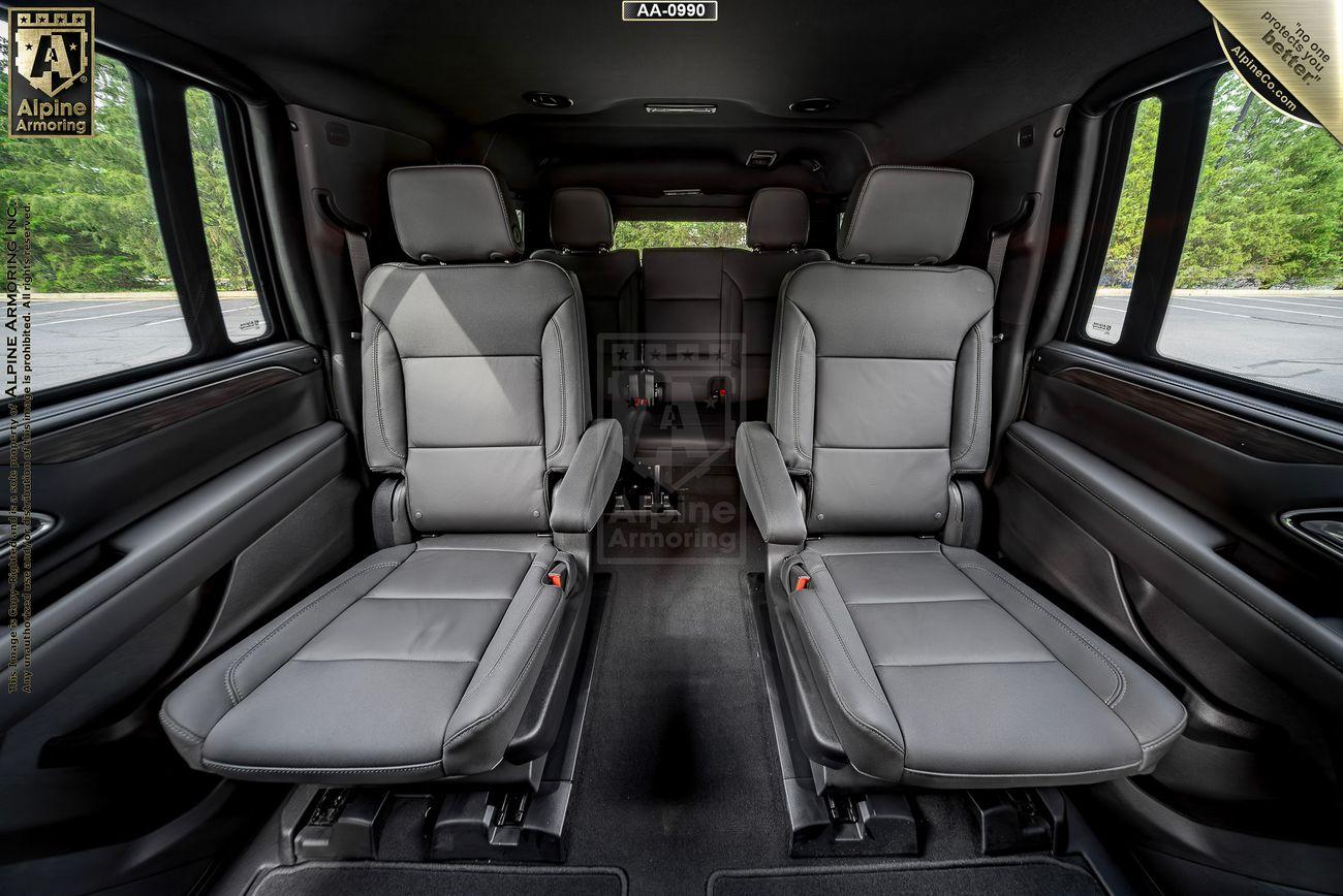 Interior view of a Chevrolet Suburban LT showing two rows of leather seats facing forward, with the front seats folded down. The windows reveal a green, wooded area outside.