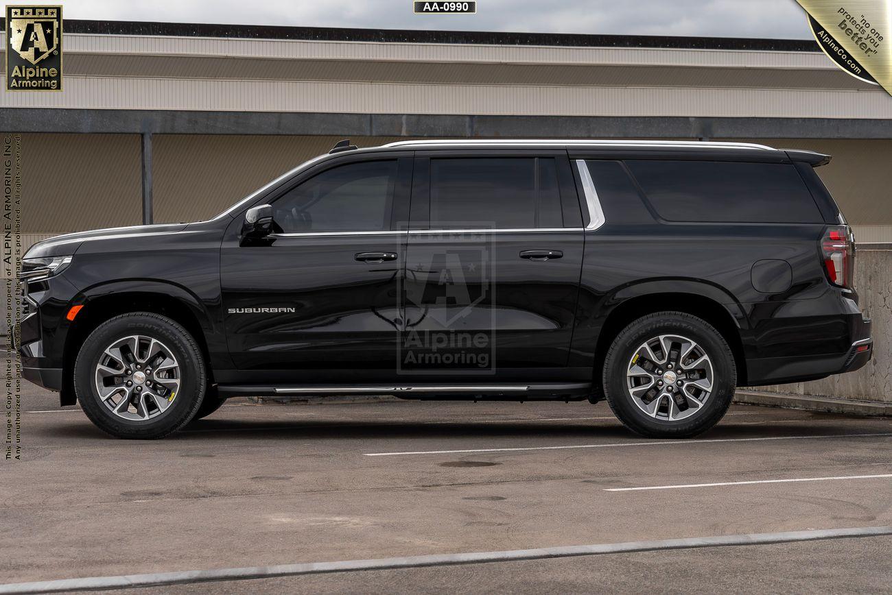 A black armored Chevrolet Suburban LT SUV labeled "Suburban" is parked in a lot near a building, displaying a logo from Alpine Armoring.