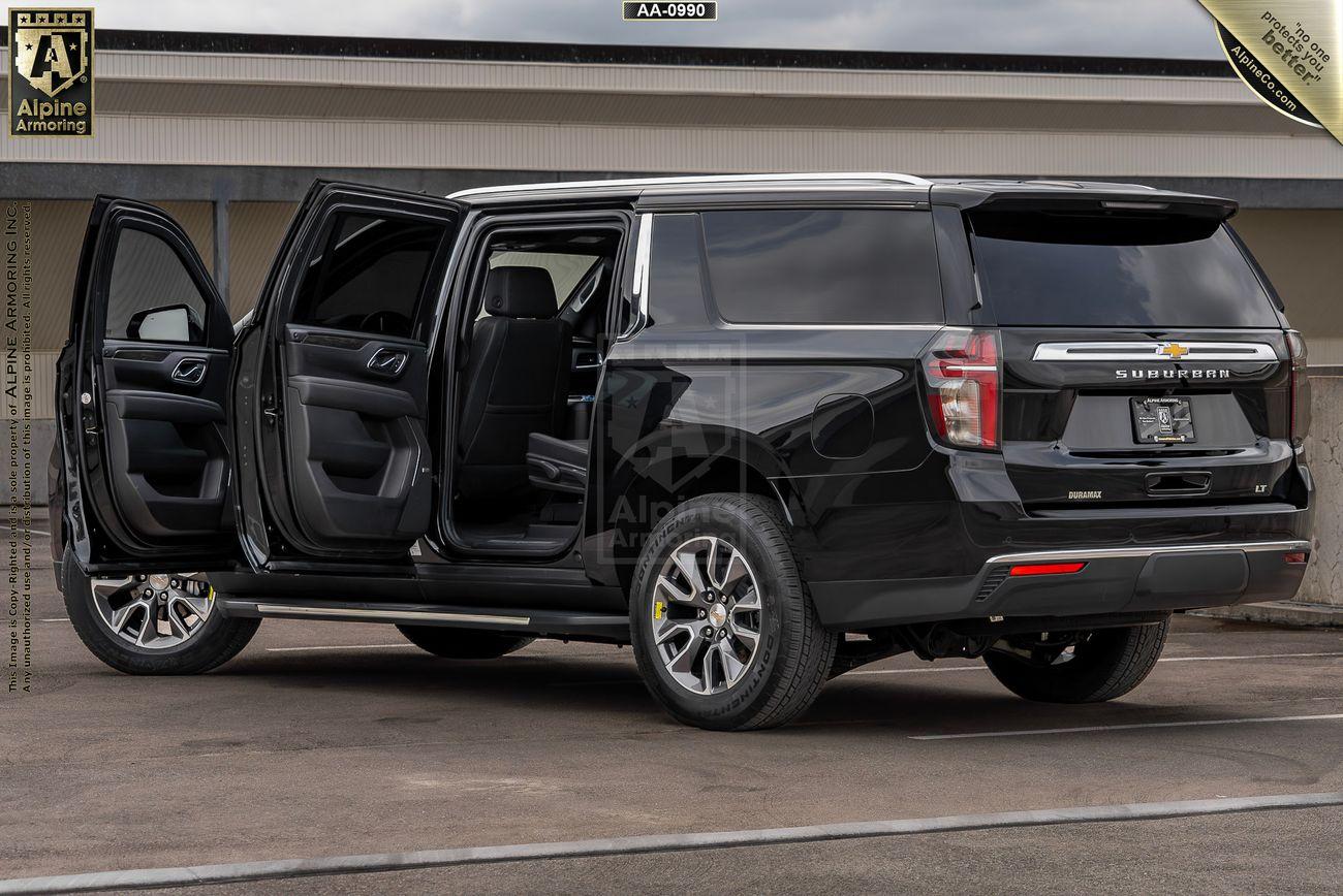 A black armored Chevrolet Suburban LT SUV with all four doors open is parked in front of a building. The vehicle has tinted windows and a silver horizontal bar on the rear.