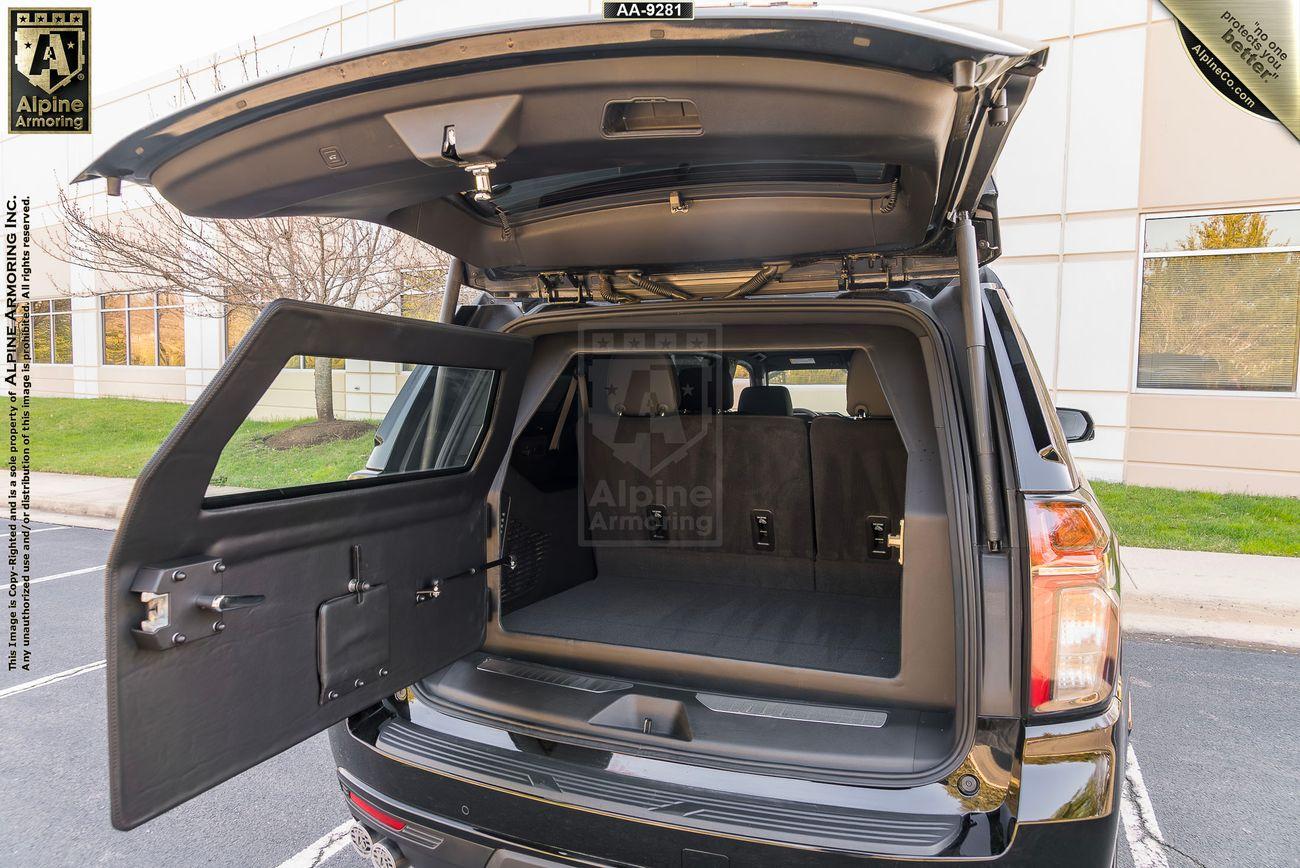 An armored Chevrolet Suburban RST SUV with the tailgate open, showcasing an installed rear gun storage and organization system. The vehicle is parked in a lot with a building in the background.