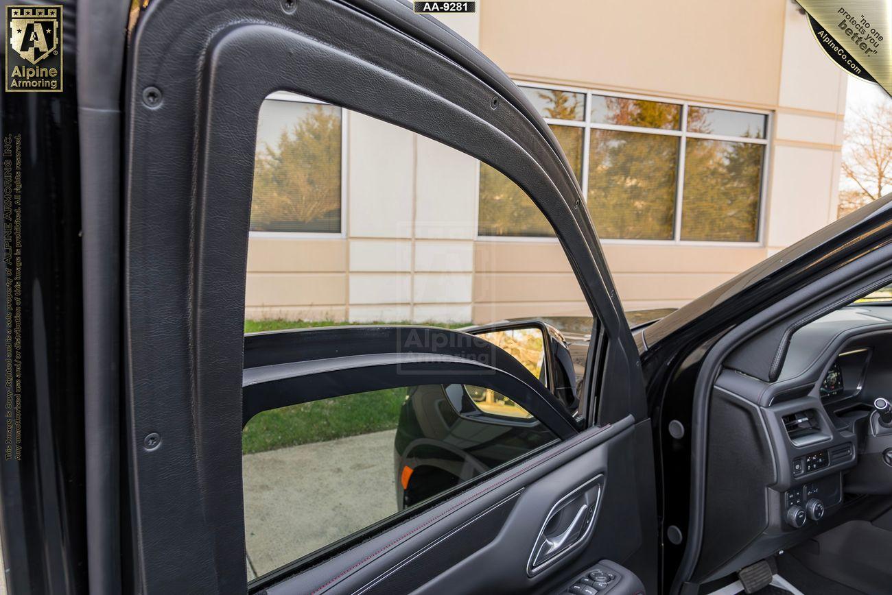 Open door of an armored Chevrolet Suburban RST SUV with visible side mirror, window, and interior panel. A building with windows and a lawn is in the background.