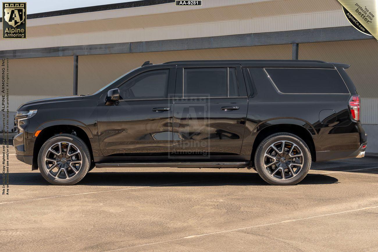 A side view of a black bulletproof Chevrolet Suburban RST SUV parked outdoors. The vehicle has dark tinted windows and is positioned in front of a large building with a beige and white facade.