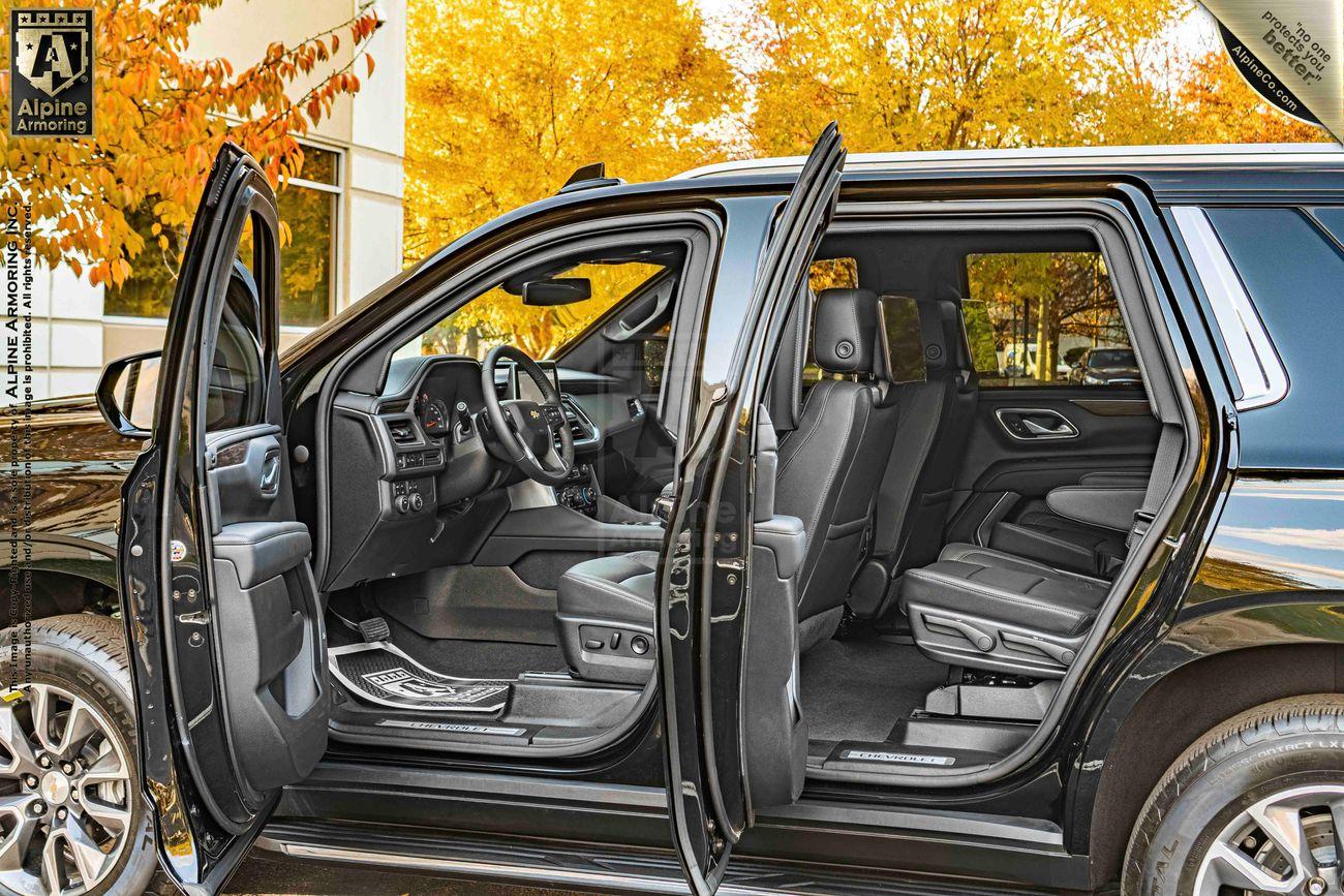 A black, bulletproof Chevrolet Tahoe SUV is shown with the front and rear doors open, revealing a spacious interior with leather seats. The background features autumn foliage.