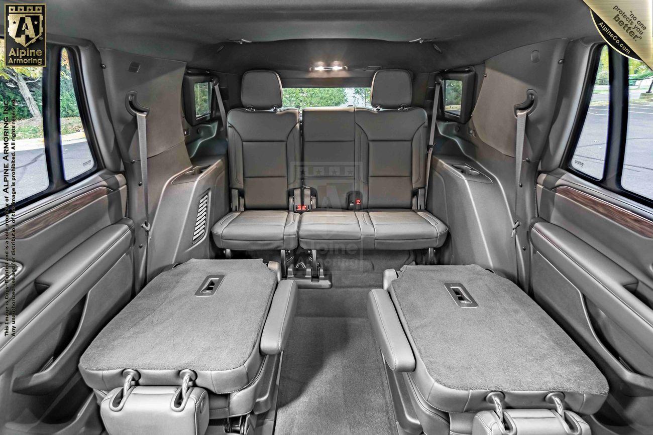 Interior of a large, luxury, armored Chevrolet Tahoe SUV with grey leather seats; second row folded flat, third row upright. Spacious cabin with light entering from rear windows. Beige trim and overhead lighting visible.