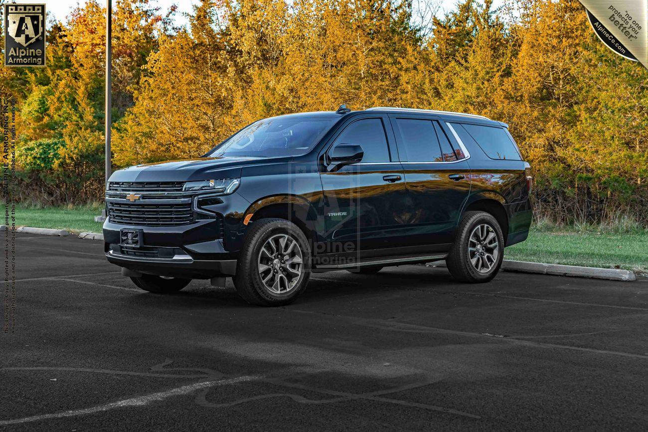 An armored black Chevrolet Tahoe SUV is parked on an asphalt surface in front of a backdrop of autumn foliage and trees.