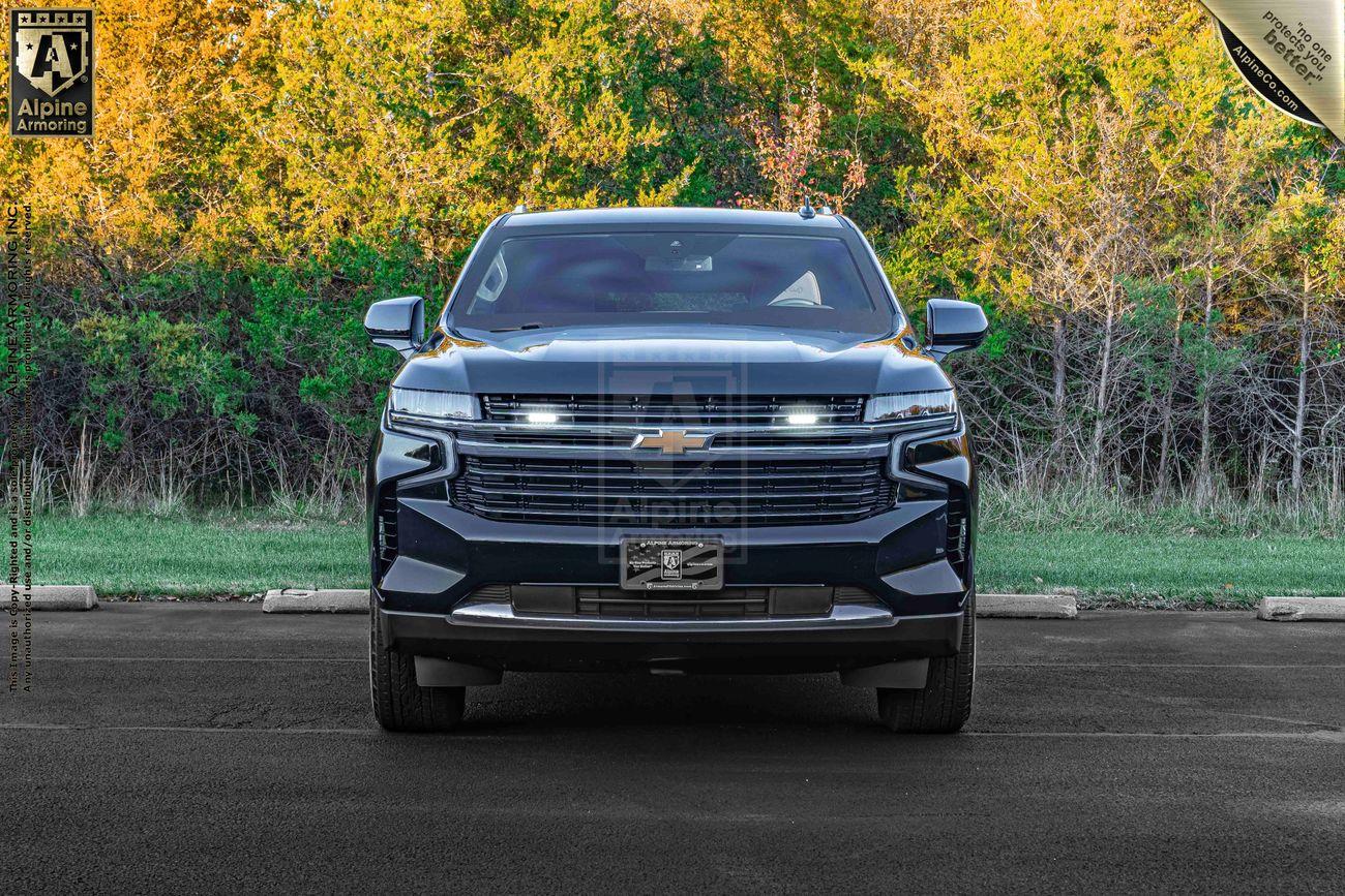 Front view of an armored black Chevrolet Tahoe SUV parked on a paved surface, with a backdrop of green trees and bushes. A dealership license plate is visible.