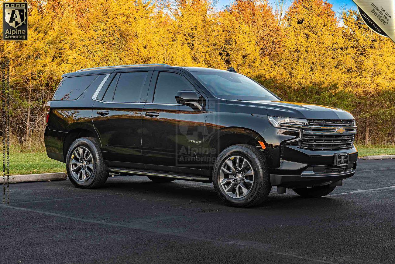 A black armored Chevrolet Tahoe SUV is parked on a paved road with autumnal trees in the background. The SUV has tinted windows and alloy wheels.