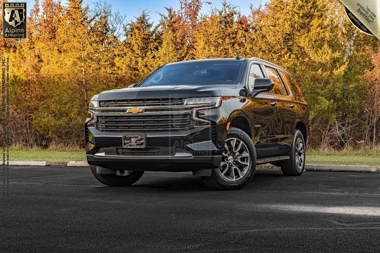 A black Chevrolet Tahoe SUV is parked on a paved surface, in front of fall-colored trees. The vehicle has Alpine branding on the windshield and a dealership plate.